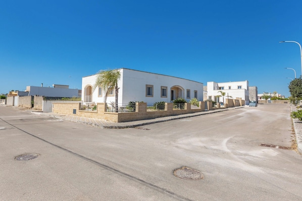 A street view with a white building featuring a central arched entrance and a small garden area.