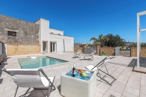 A poolside area with lounge chairs, a table with food and drink, and a building with a stone facade.