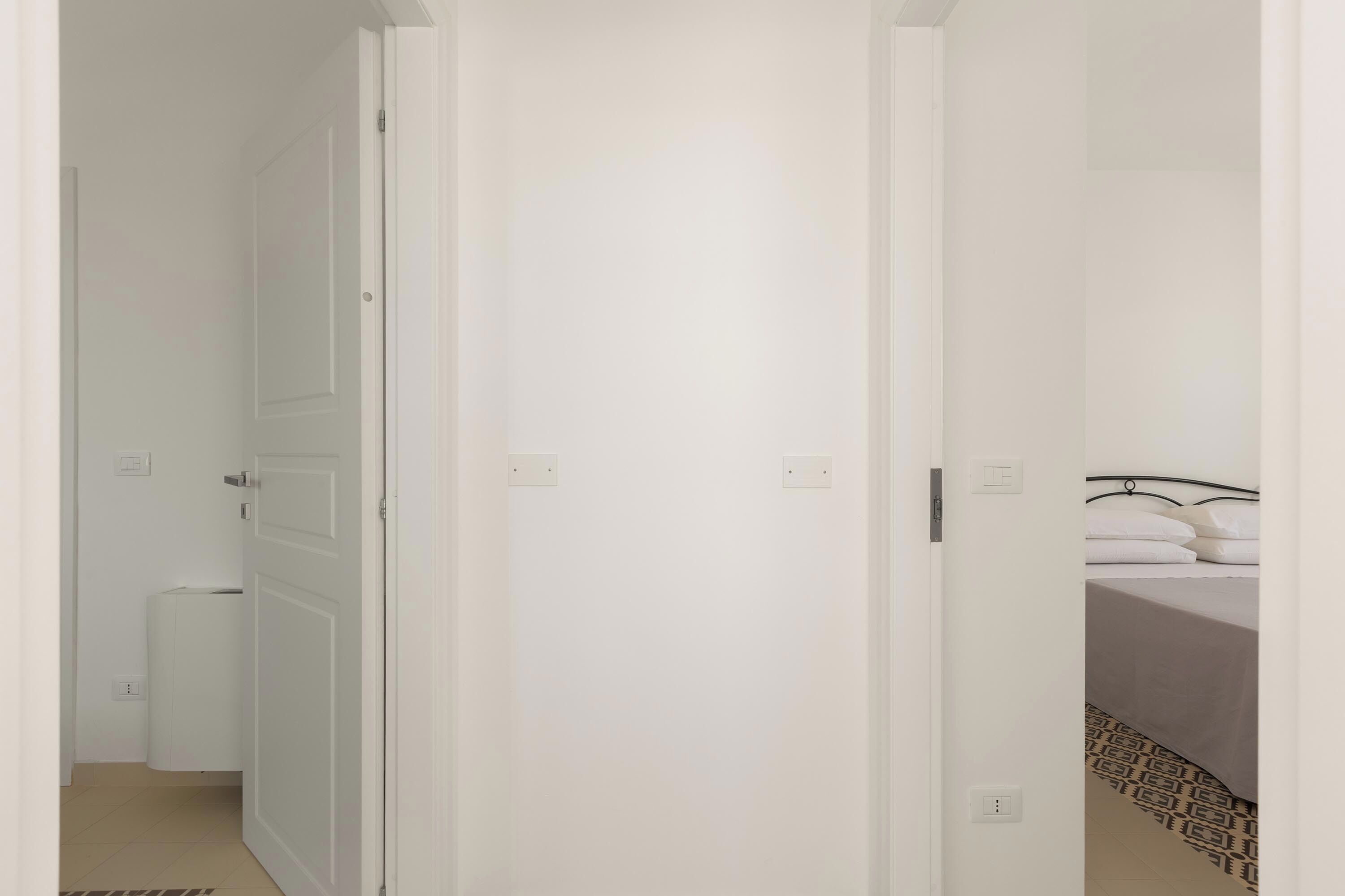 Ein schmaler Flur, der zu einem Zimmer mit einem Bett, einem Heizkörper und einer Tür führt.