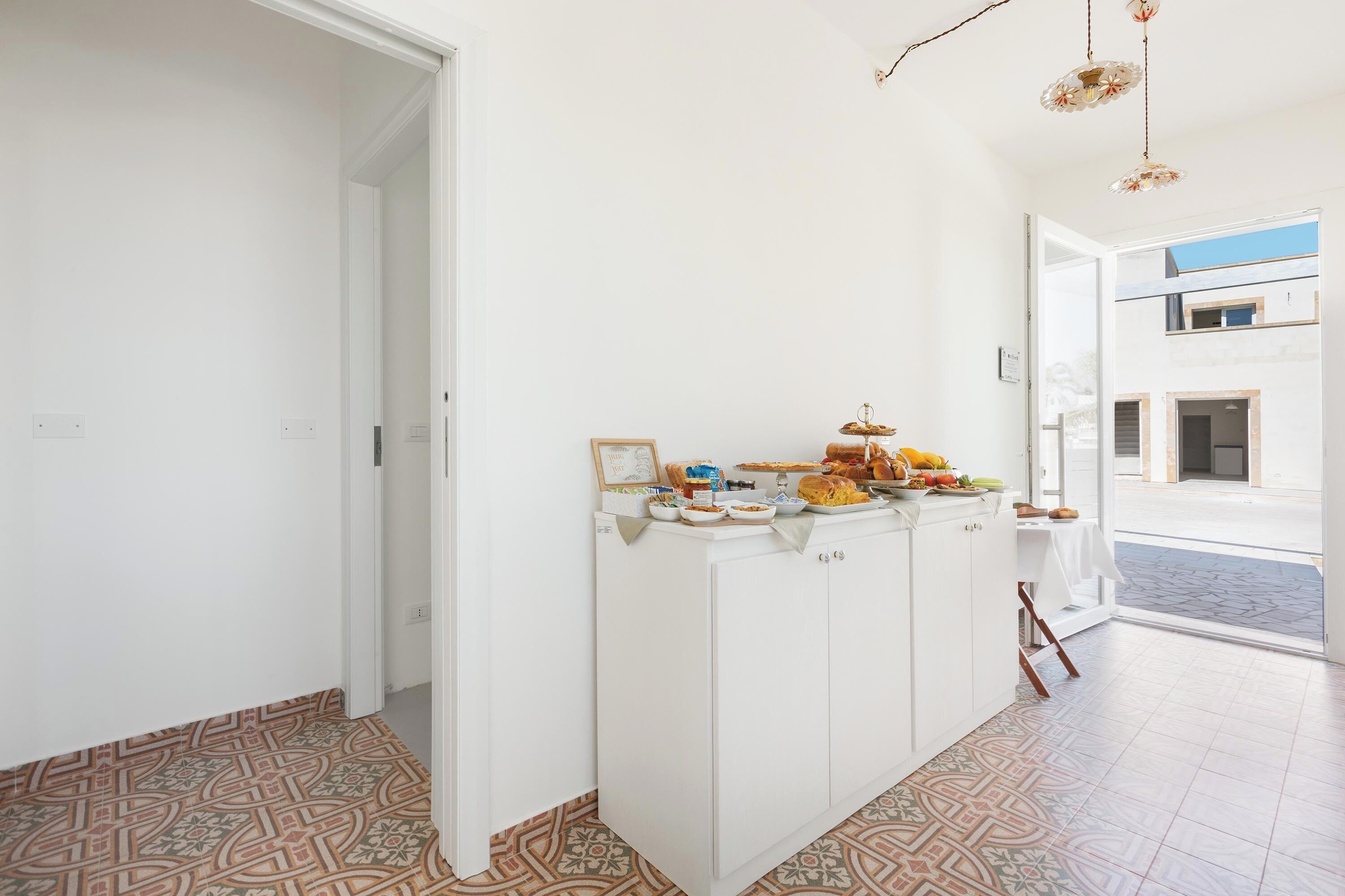 Un corridoio con pavimento decorato, un armadietto bianco pieno di alimenti e vista su uno spazio esterno.