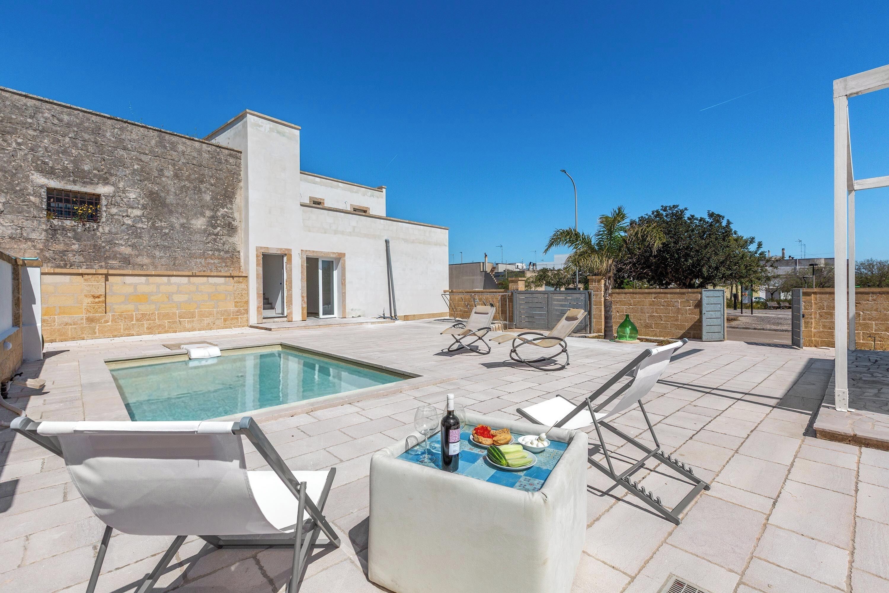 Zona piscina con poltrona relax, tavolo con cibo e bevande, e un edificio con facciata in pietra.