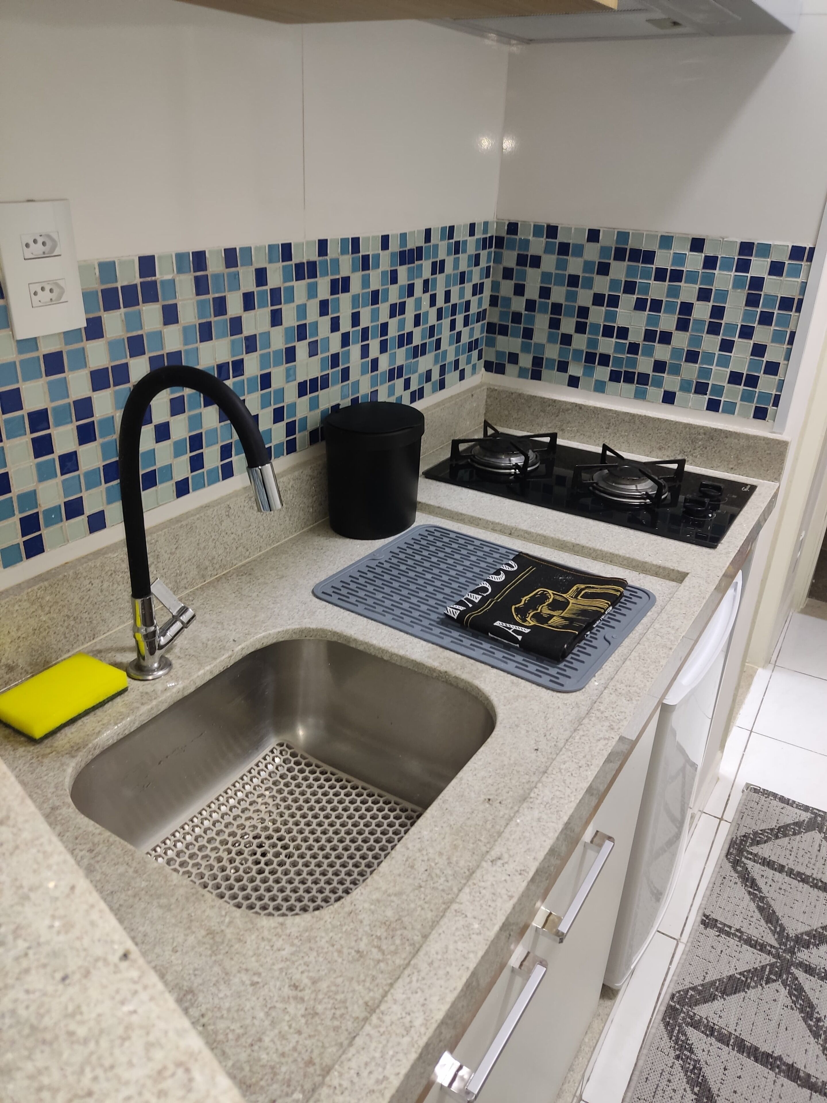 A kitchen with a stainless steel sink, a black trash bin, a gas stove, and a blue-tiled backsplash.