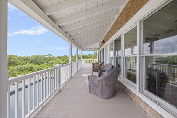 A covered outdoor deck with wicker furniture, large windows, and a view of greenery.
