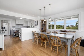 A modern kitchen with white cabinetry, a central island with bar stools, pendant lighting, and a view of greenery outside.