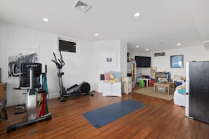 A spacious room with a wooden floor, a treadmill, an elliptical, a desk with a chair, a TV, and a refrigerator.