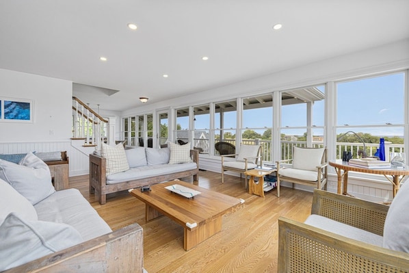 A spacious living room with a wooden floor, white furniture, and large windows offering a view of the outdoors.