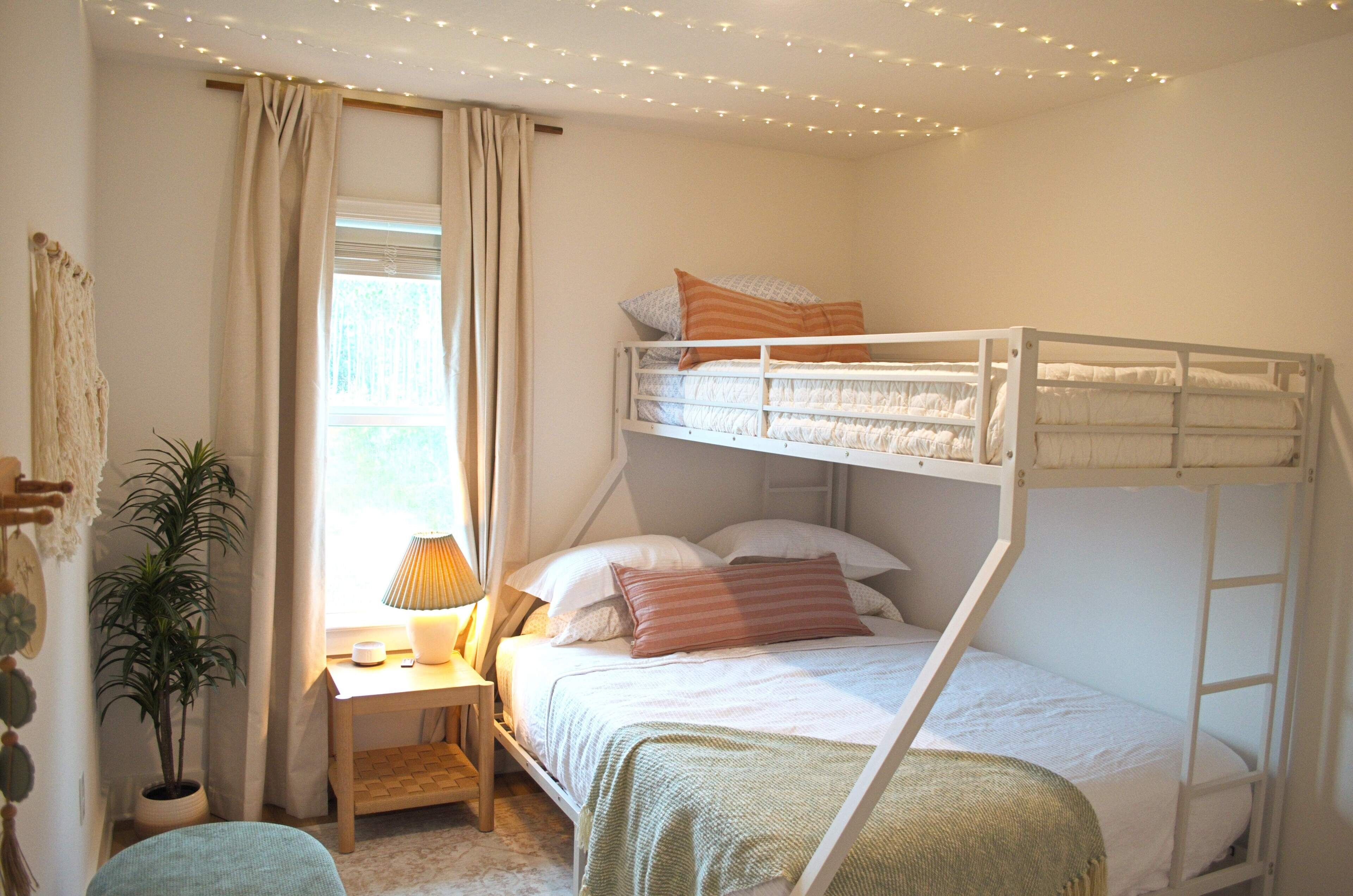 A bunk bed with a bedside table and lamp, a potted plant, and a window with curtains.