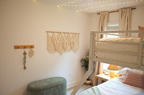 Een stapelbed met nachtkastje, een klein krukje, wanddecoratie van macramé en een lichtslingering.