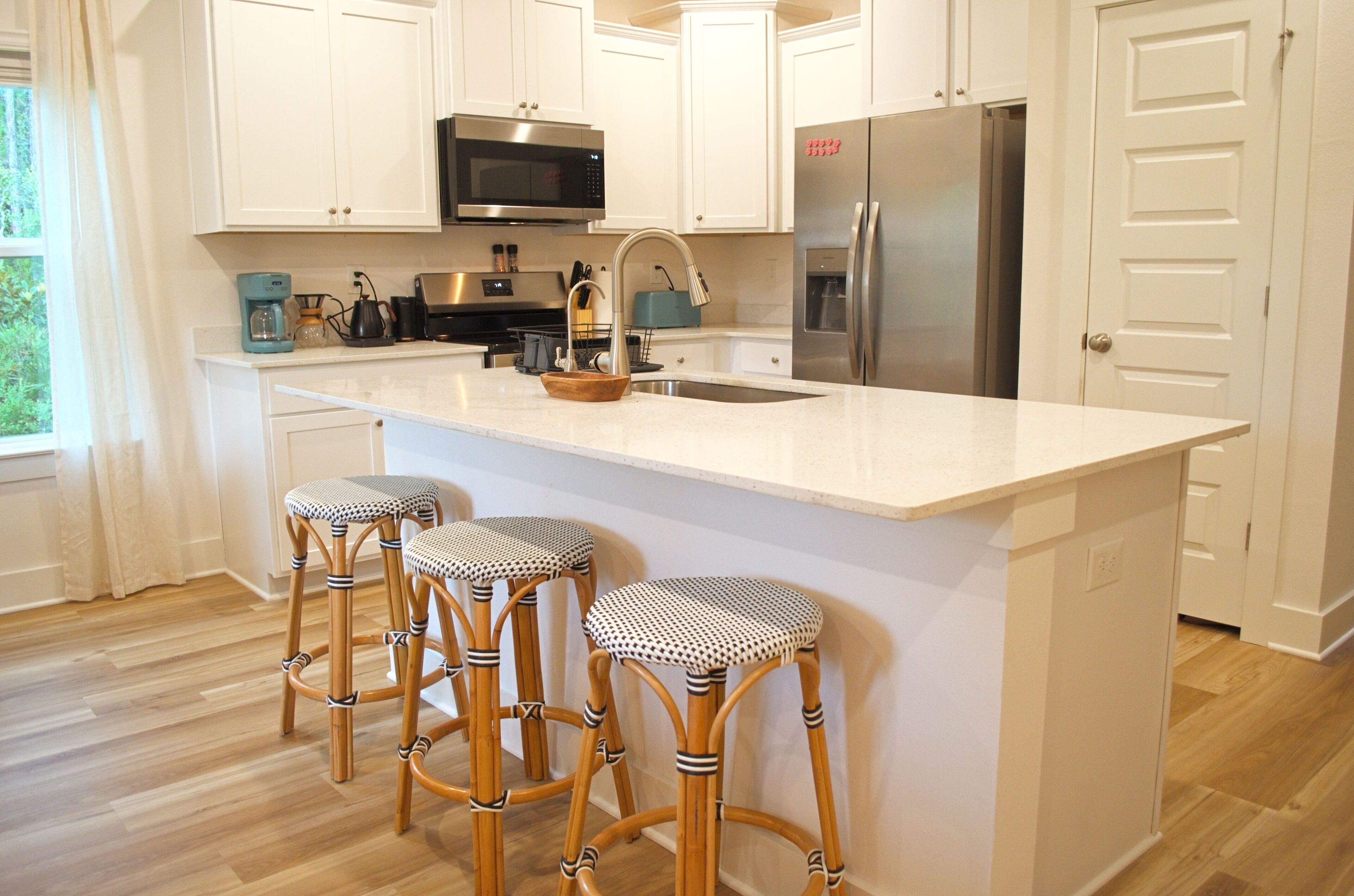 A kitchen with white cabinets, a stainless steel refrigerator, a microwave, and a sink. There are three bar stools with a checkered seat pattern.