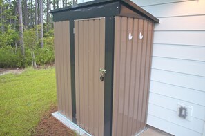 A brown metal shed with a black roof, a lock, and a small window, located next to a house with white siding. - Boho Coastal Retreat - Bay + 30A Beaches - Tesla Charger - Sleeps 7 (Santa Rosa Beach)
