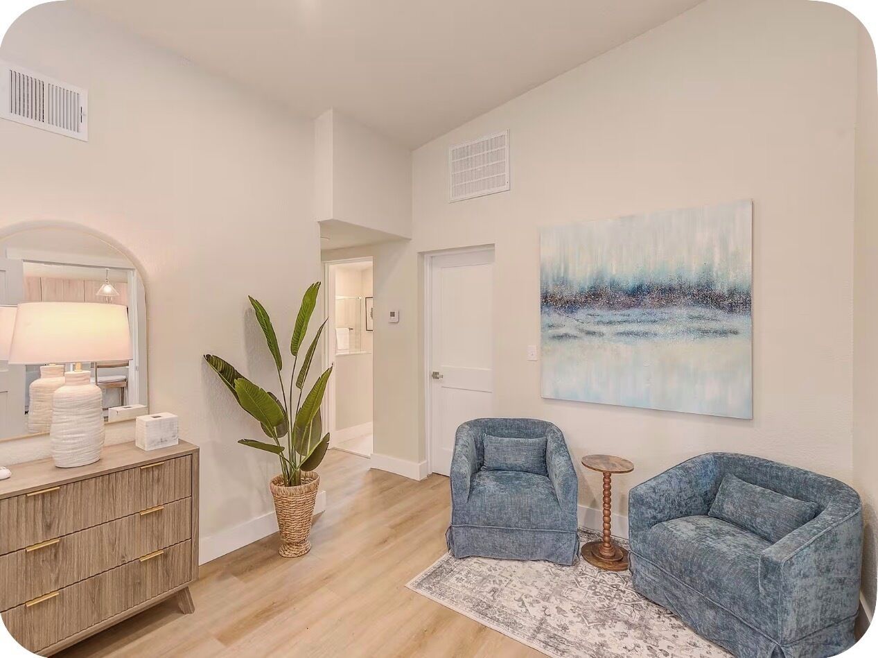 A modern living room with a wooden floor, a large potted plant, two blue armchairs, a small round table, and a painting on the wall.