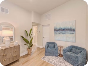 Uma sala de estar moderna com piso de madeira, uma grande planta em vaso, duas poltronas azuis, uma pequena mesa redonda e um quadro na parede.