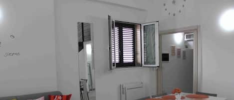 Una sala moderna con un sofá gris, una mesa de comedor con cuatro sillas y una ventana con persianas.