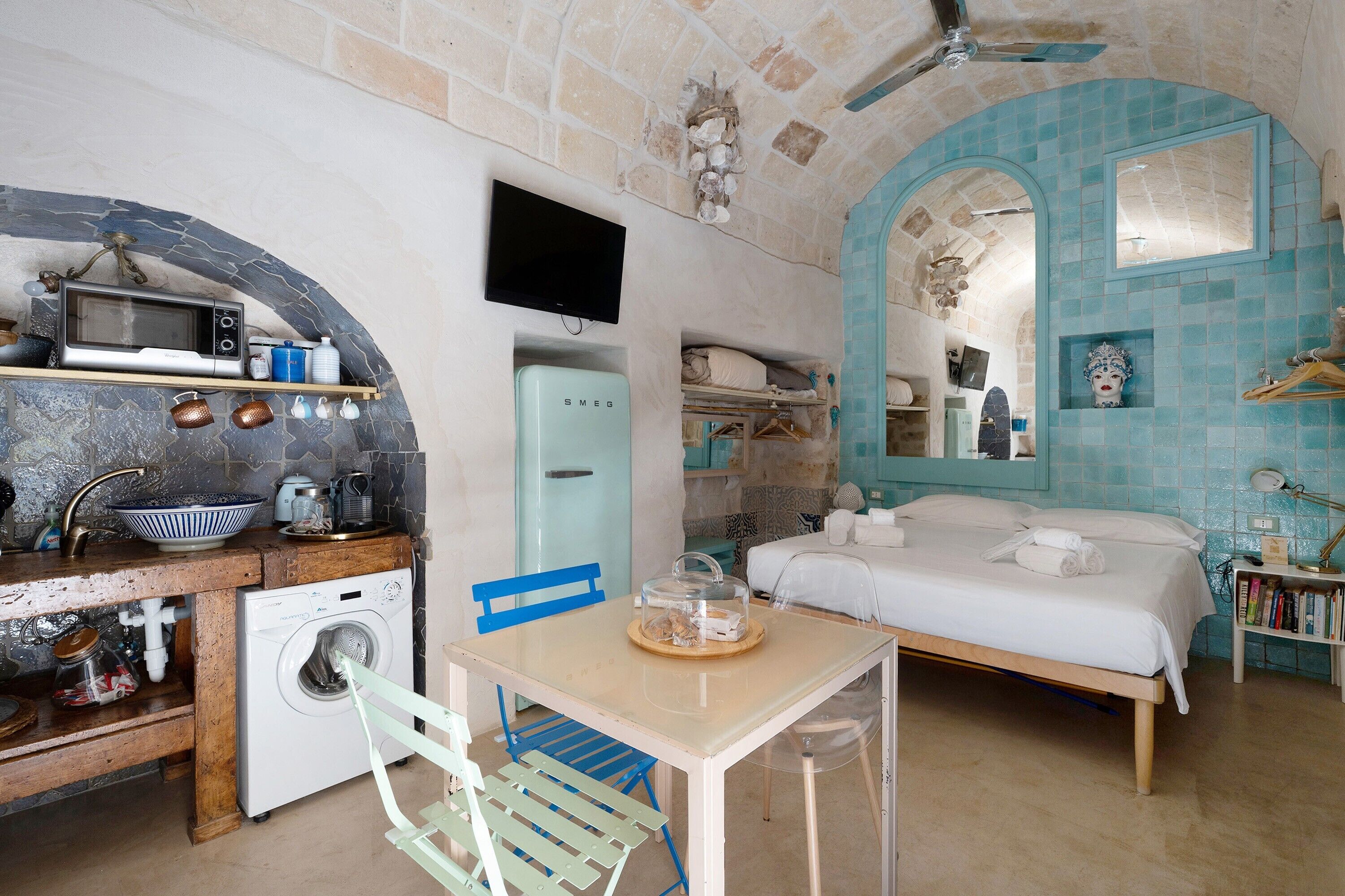 Une pièce compacte comprenant un coin cuisine, un lit, une télévision et une salle de bain avec un mur recouvert de carreaux bleus.