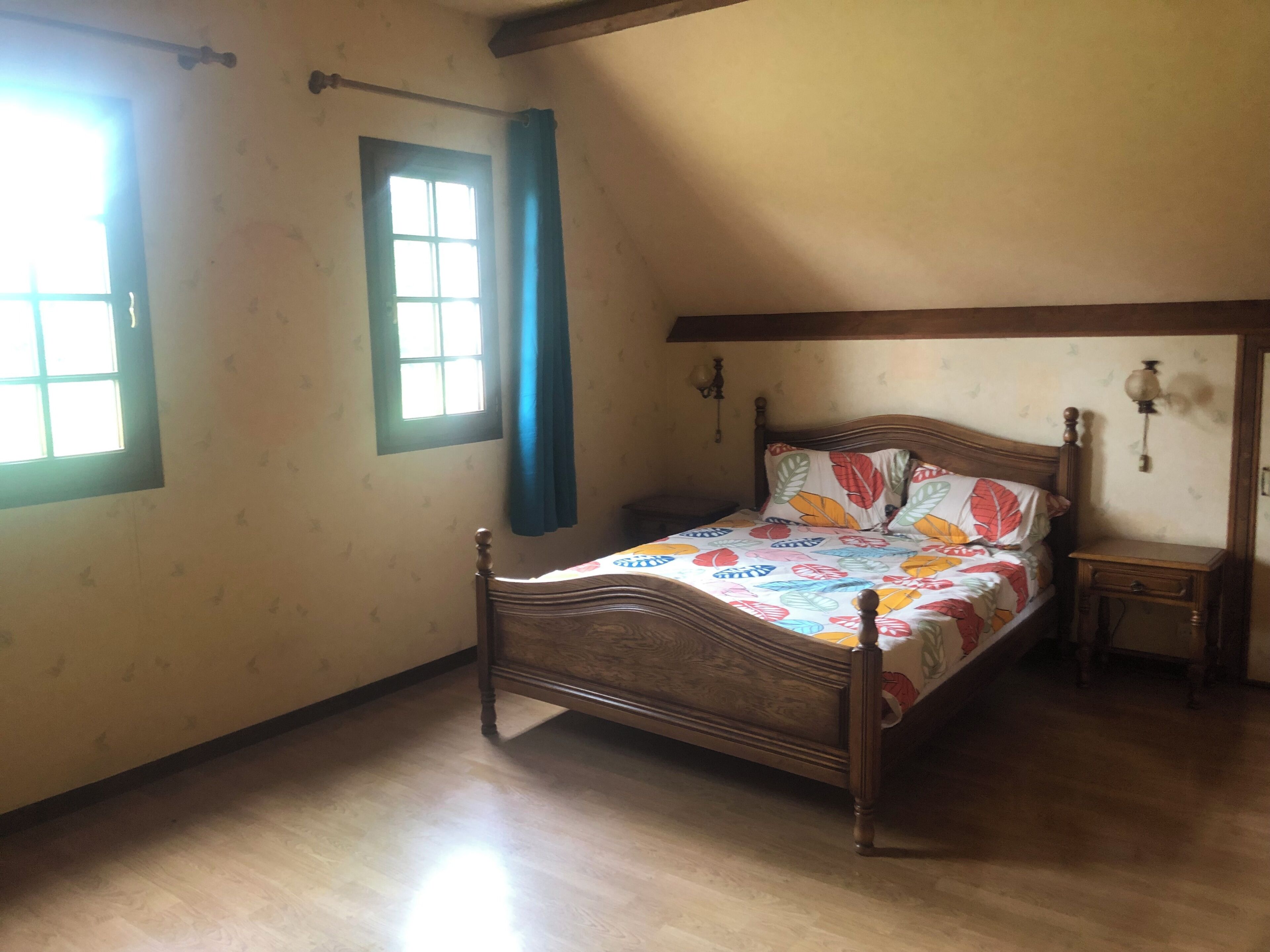 A bedroom with a wooden bed, a nightstand, a window with curtains, and a wall-mounted light fixture.