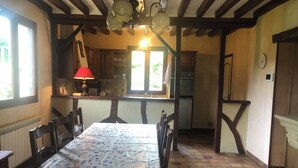 Un coin repas avec une table dressée pour quatre personnes, des poutres en bois au plafond, une cuisine en arrière-plan et une fenêtre laissant entrer la lumière naturelle.