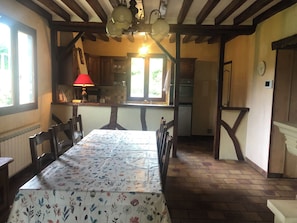 Ein Essbereich mit Tisch für vier Personen, Holzbalken an der Decke, eine Küche im Hintergrund und ein Fenster mit Tageslicht.
