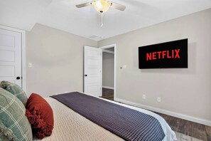 A bedroom with a bed, pillows, a wall-mounted TV displaying 'NETFLIX', and a ceiling fan.