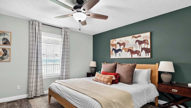 Een slaapkamer met een bed, een plafondventilator, een raam met zonwering, een schilderij van paarden en een nachtkastje met een lamp.
