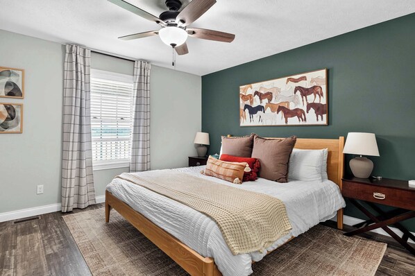 A bedroom with a bed, a ceiling fan, a window with blinds, a painting of horses, and a nightstand with a lamp.