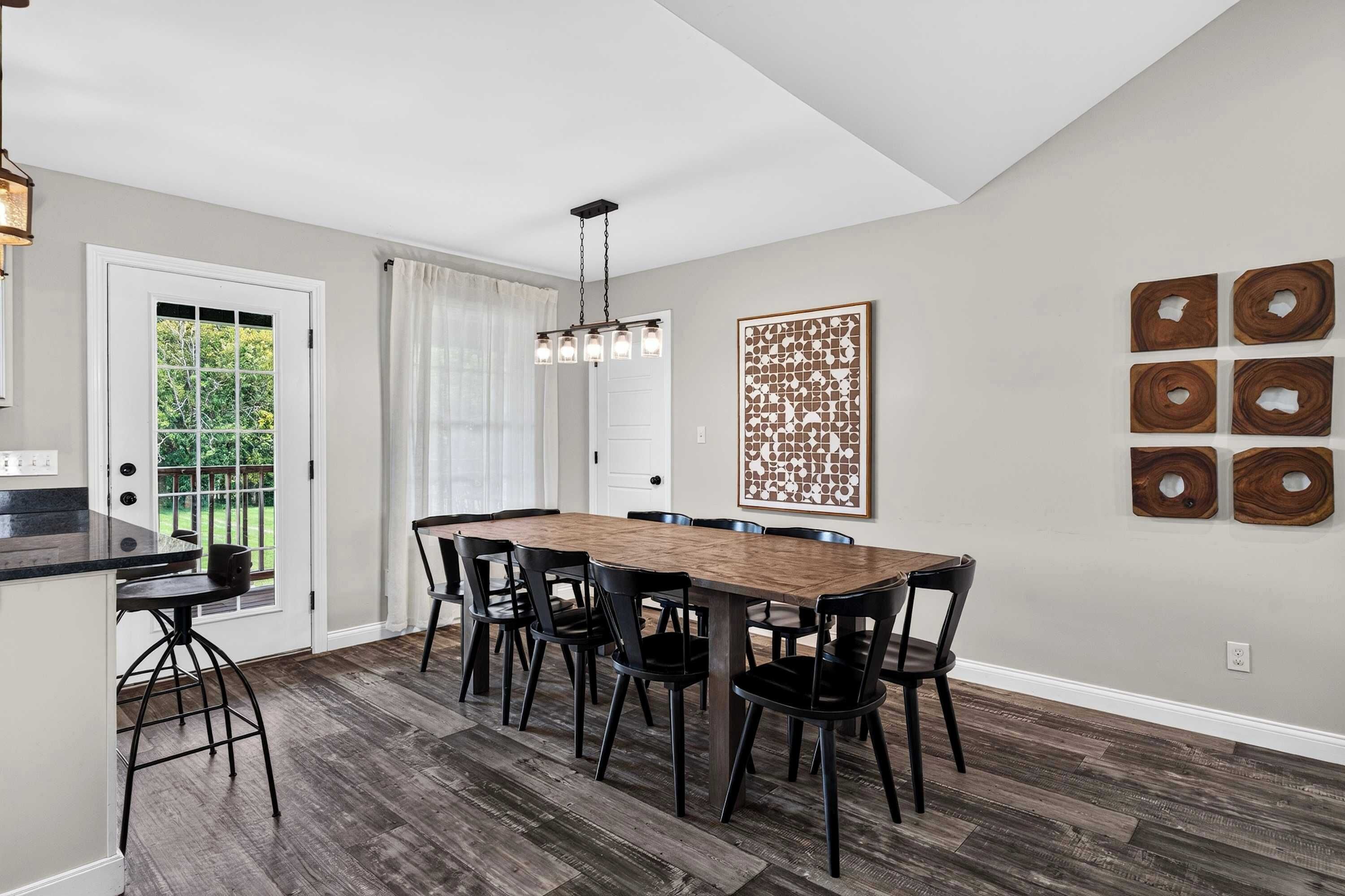 Un espace repas comprenant une table et des chaises en bois, un luminaire suspendu et une vue sur l’extérieur par une porte.