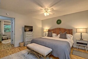 Quarto com cama, luminárias de cabeceira, televisão, ventilador de teto e uma porta que dá para outro espaço.