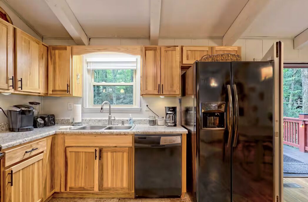 A kitchen with wooden cabinets, a stainless steel refrigerator, a dishwasher, and a window with a view of greenery.