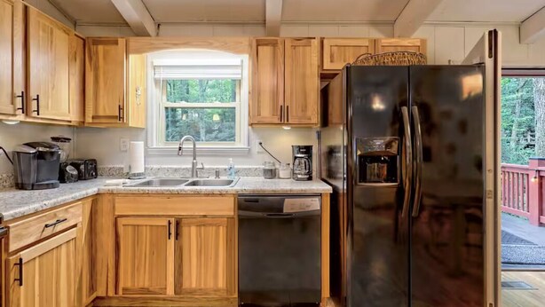 A kitchen with wooden cabinets, a stainless steel refrigerator, a dishwasher, and a window with a view of greenery. - Little Creek Cabin — Sky Valley (Dillard)