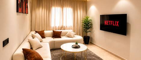 A modern living room with a sectional sofa, a coffee table, a potted plant, and a wall-mounted TV displaying the Netflix logo.