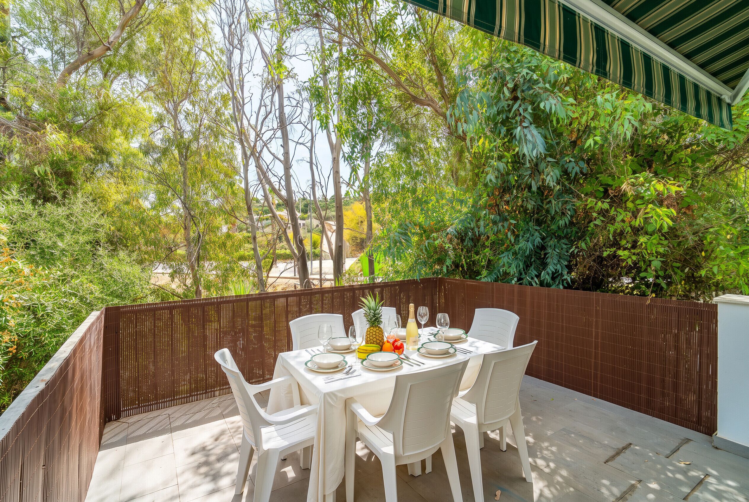 Un espace extérieur aménagé pour les repas, avec une table dressée pour quatre personnes, entouré d’une clôture en bois et donnant sur un jardin planté d’arbres et d’arbustes.