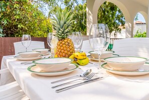 Una mesa de comedor al aire libre dispuesta con platos blancos, cuencos con borde verde y copas de vino transparentes, con una piña como centro de mesa y una pequeña figurita amarilla.