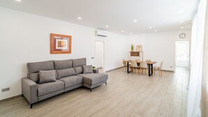 A modern living room with a grey sectional sofa, a dining area with a wooden table and chairs, and a white wall with a framed picture and an air conditioning unit. - Cubo's Afrodita House / urban / terrace / air conditioning / free wifi (Alhaurín el Grande)