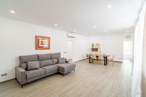 Ein modernes Wohnzimmer mit einem grauen Ecksofa, einem Essbereich mit Holztisch und -stühlen sowie einer weißen Wand mit einem gerahmten Bild und einer Klimaanlage.