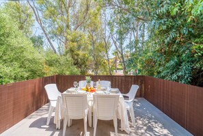 An outdoor dining area with a table set for four, surrounded by white plastic chairs, with a view of trees and greenery.