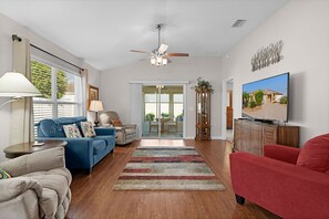 A living room with a blue sofa, a red armchair, a wooden cabinet, a television, and a ceiling fan. - Private Courtyard Villa with All-Weather Lanai Near Brownwood (The Villages)