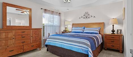 A bedroom with a bed, a dresser, a nightstand, a ceiling fan, and a window with curtains.