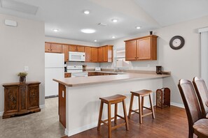 A kitchen with wooden cabinets, a white refrigerator, a microwave, a stove, and a sink. There are two wooden stools and a wooden chair. - Private Courtyard Villa with All-Weather Lanai Near Brownwood (The Villages)