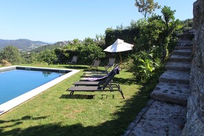 Área da piscina com espreguiçadeiras e um grande guarda-sol, rodeada por vegetação e vista para colinas ao longe.