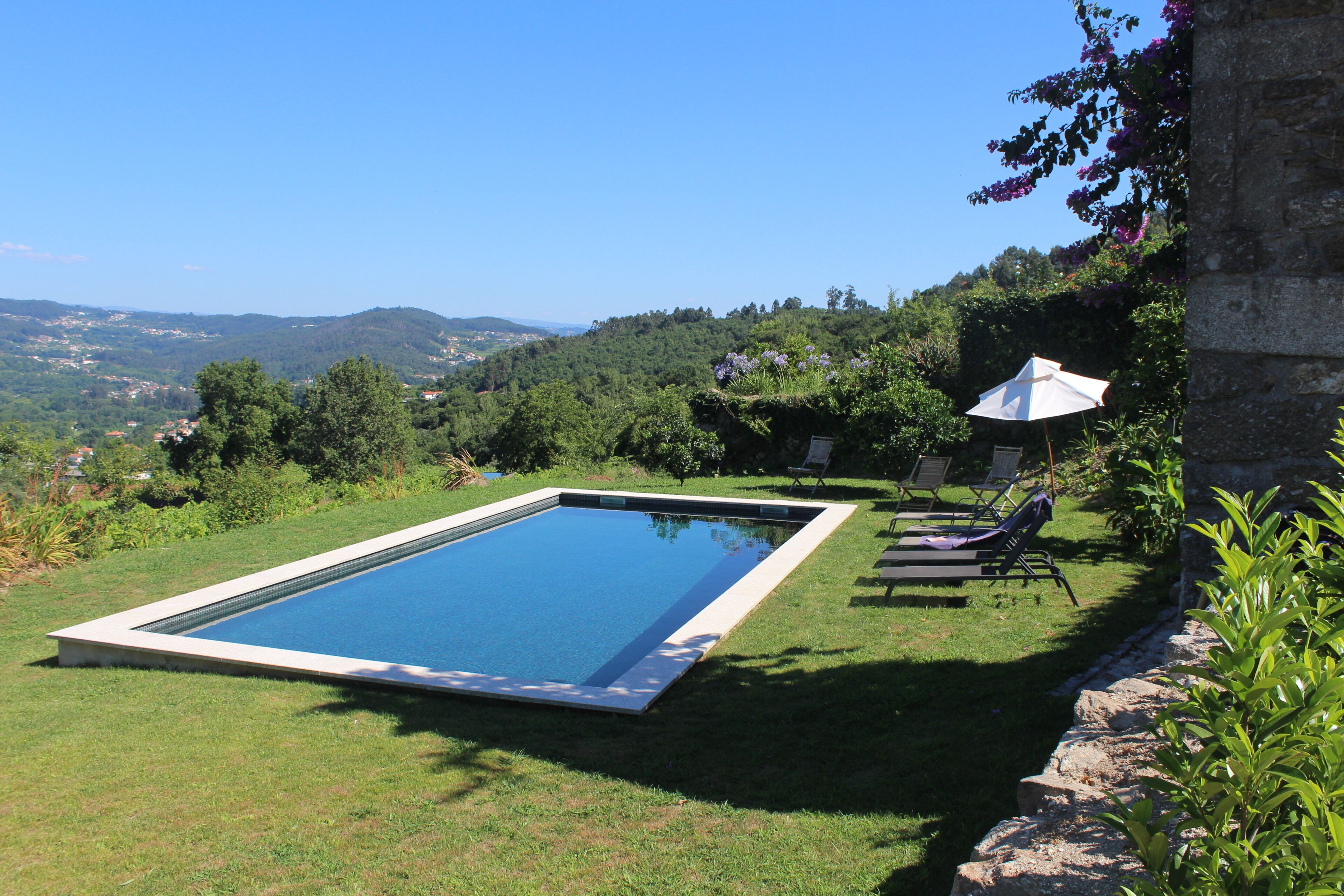 A rectangular swimming pool with a clear blue water surface, surrounded by a grassy area with a few lounge chairs and an umbrella.