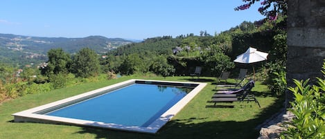 A rectangular swimming pool with a clear blue water surface, surrounded by a grassy area with a few lounge chairs and an umbrella.