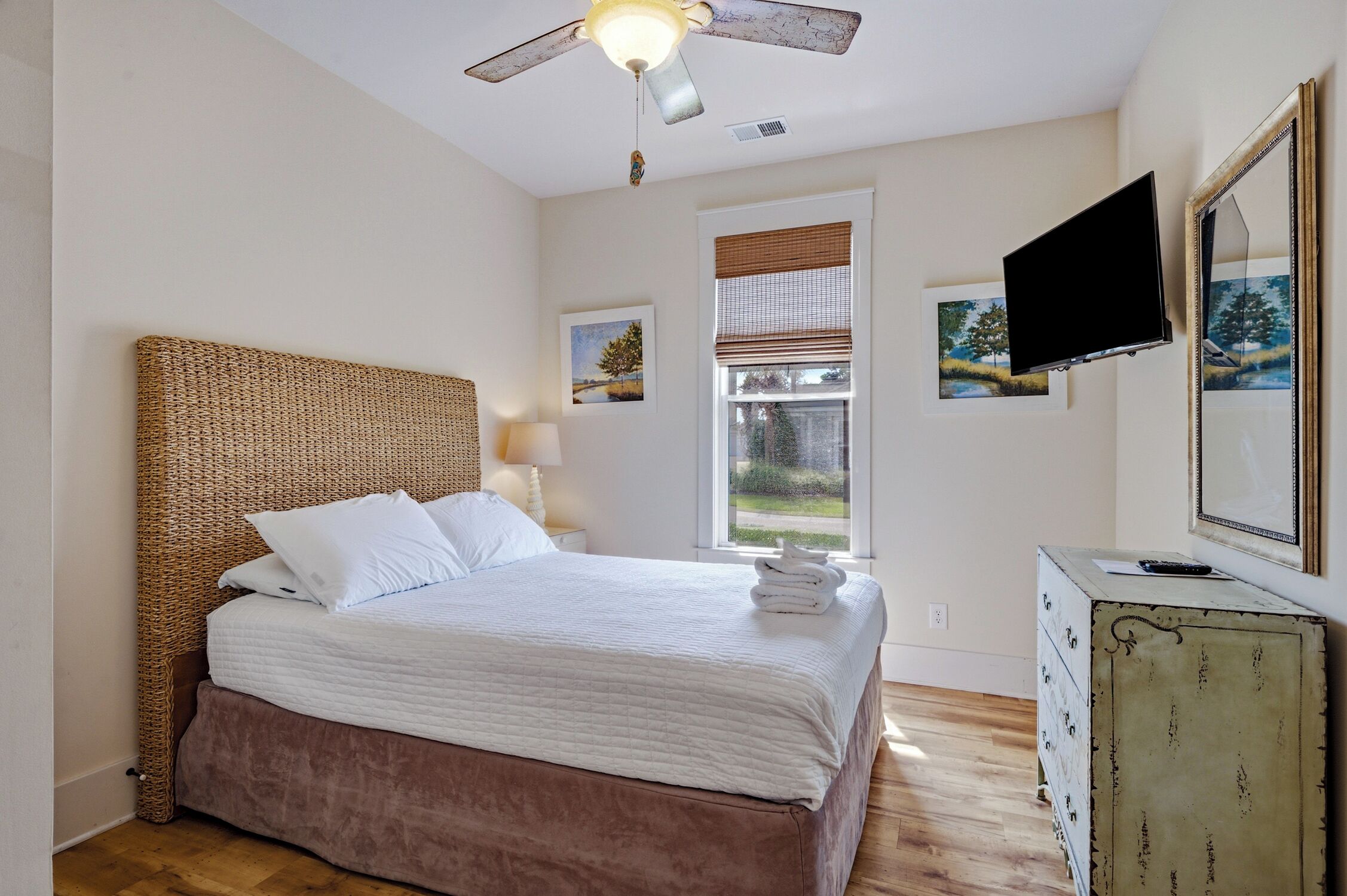 A bedroom with a bed, a ceiling fan, a window with blinds, a TV, and artwork on the wall.
