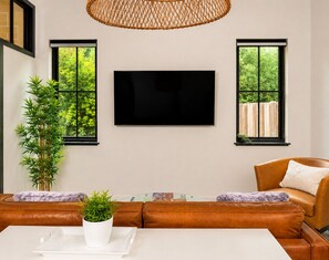 Un salon doté d’un téléviseur à écran plat fixé au mur, d’un canapé en cuir marron, d’une table basse blanche ornée d’une plante en pot et de deux fenêtres donnant sur un paysage verdoyant.