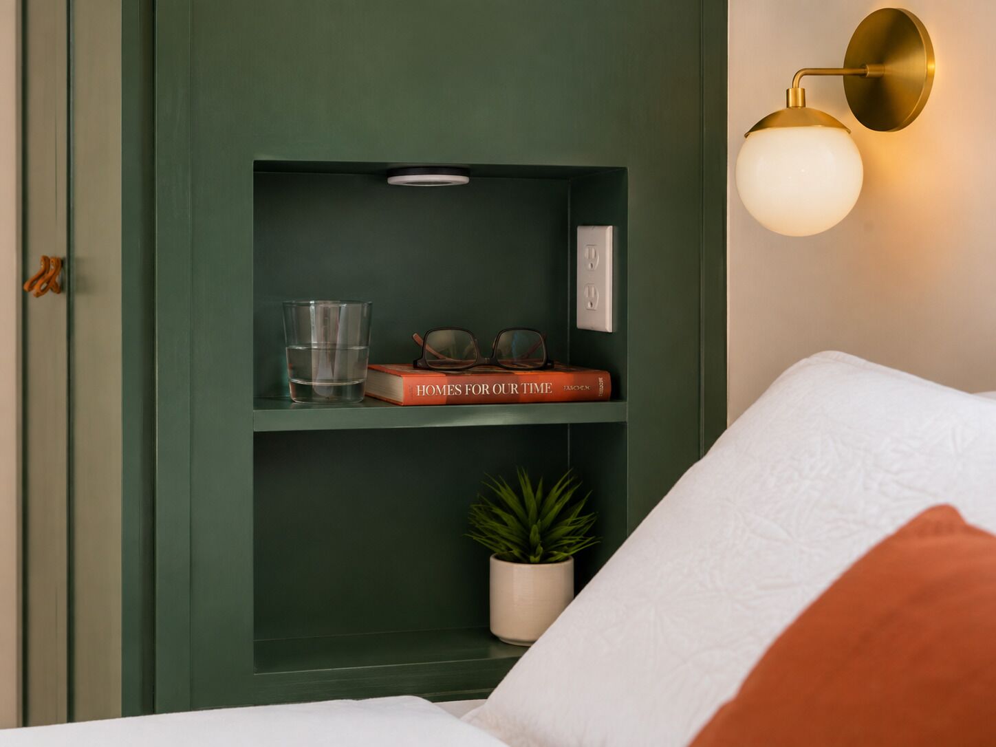 A room with a green wall, a bookshelf with books and glasses, a potted plant, and a wall-mounted light fixture.