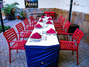 Mesa lista para una comida, con sillas rojas, un muro de piedra y una señalización que indica 'ADASA LOUNGE & SUITES' al fondo.