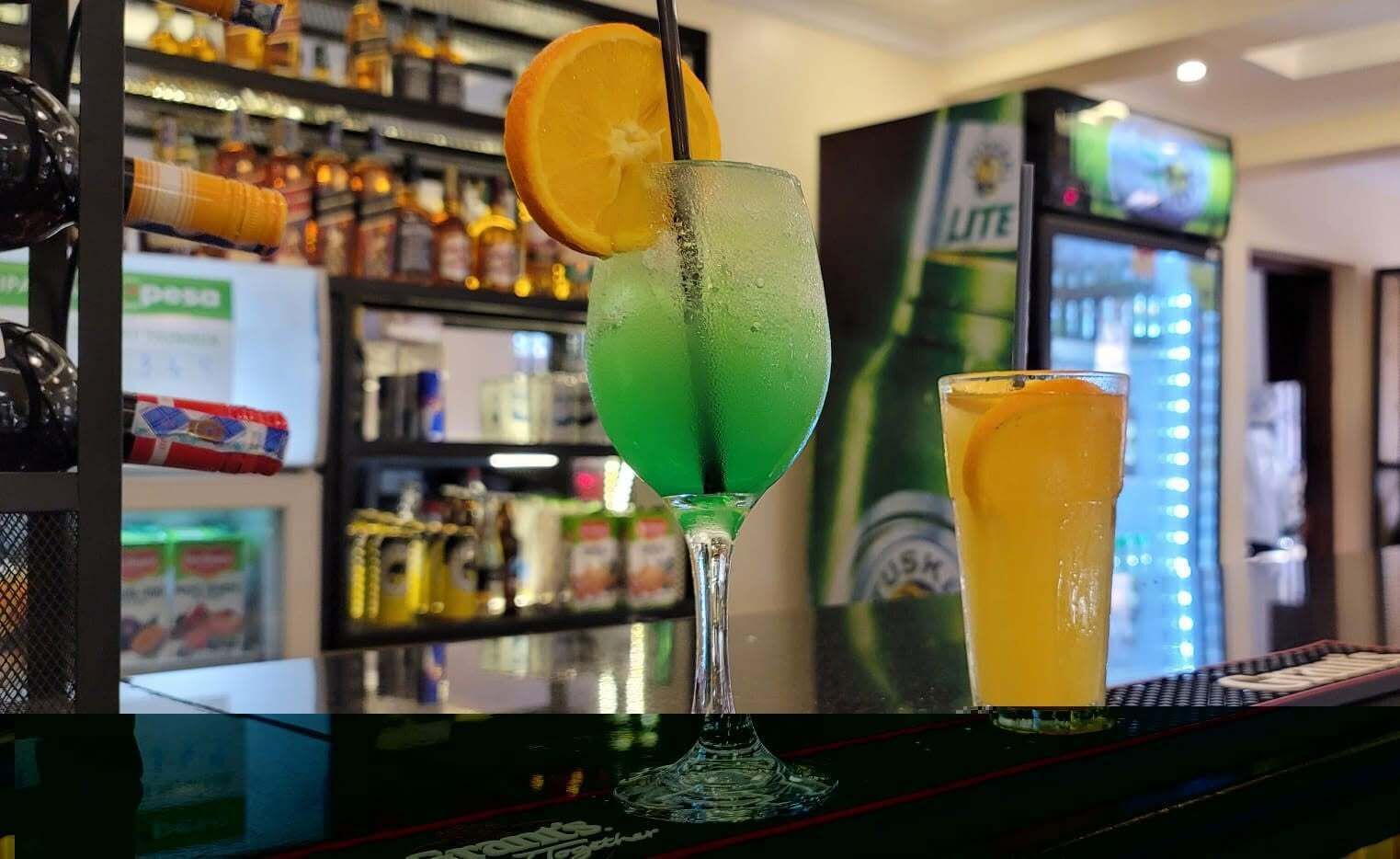 Zona de bar con un cóctel verde, un vaso alto con una rodaja de naranja y una botella de cerveza LITE al fondo.