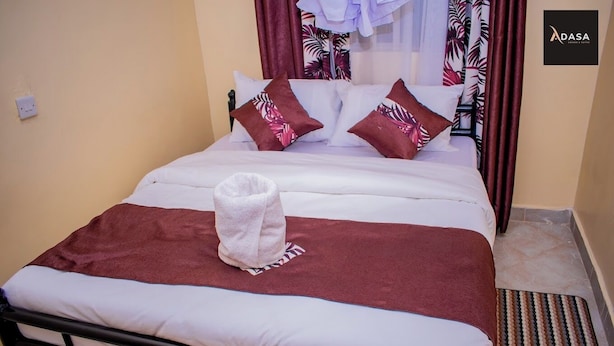 A neatly made bed with a white and brown blanket, a white towel, and two decorative pillows with a tropical leaf pattern. - Adasa Lounge and Suites (Kisumu)