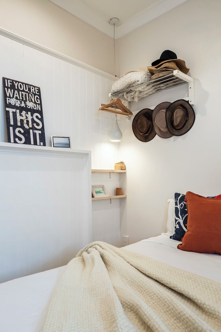 A bedroom with a bed, a wall-mounted hat rack, a shelf with hats, a framed sign, and a blanket.