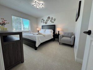 Quarto com cama, mesas de cabeceira, uma cadeira, escrivaninha e um objeto decorativo na parede.