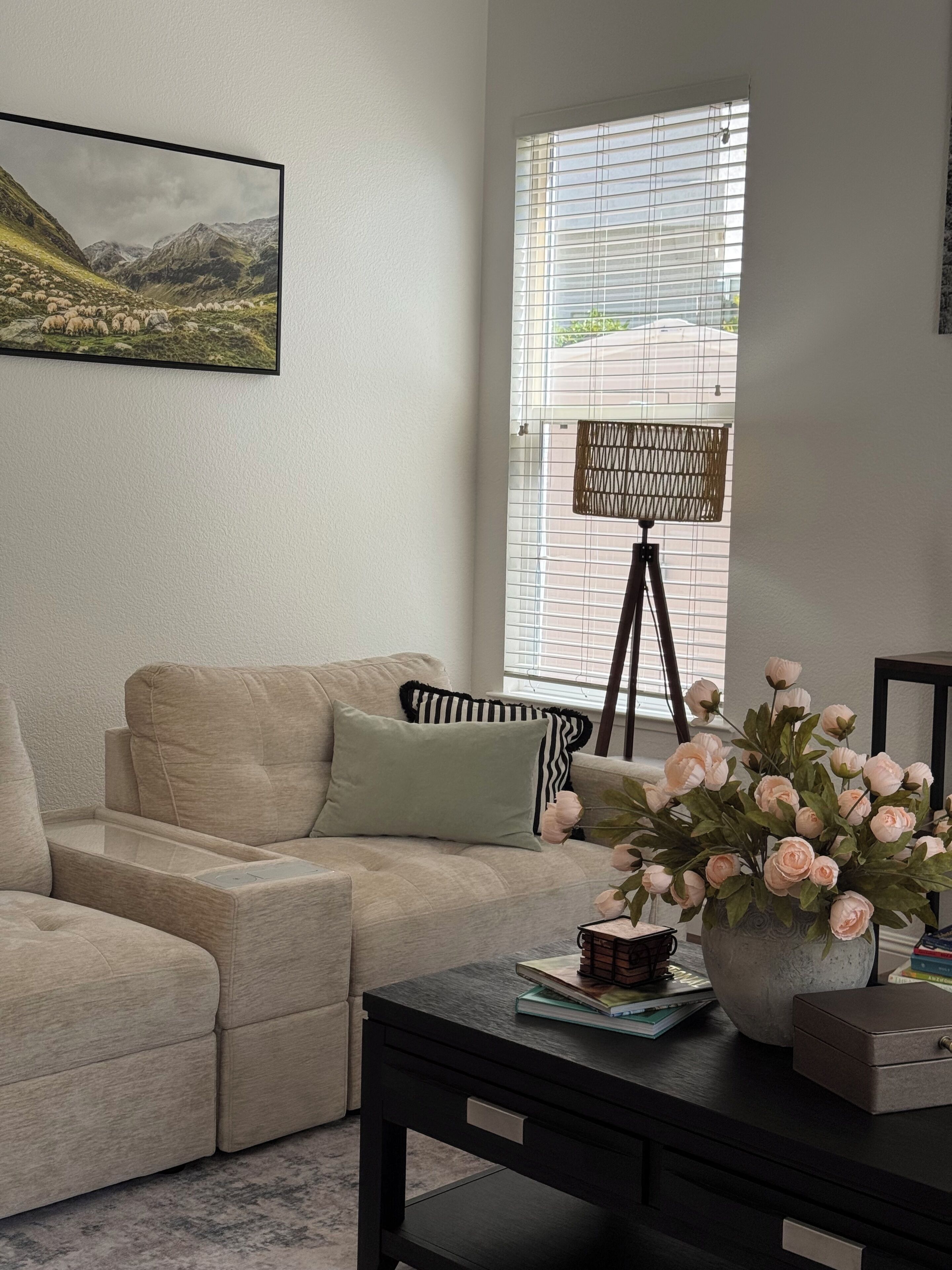 Un salon avec un canapé d’angle beige, une petite table sur laquelle est posé un vase de fleurs et une fenêtre avec des stores.