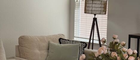 A living room with a beige sectional sofa, a small table with a vase of flowers, and a window with blinds.